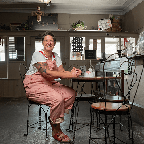 Smiling woman wearing white shirt and red overalls sitting at a table in a cafe.